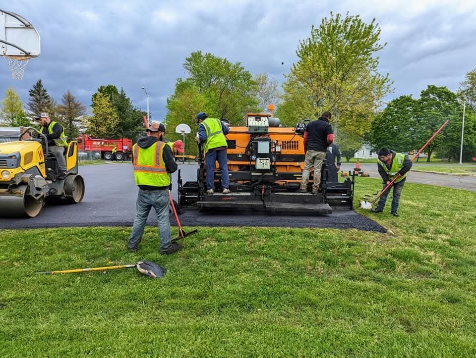 Construction crew paving asphalt near a basketball court. Workers in safety vests use machinery and tools.