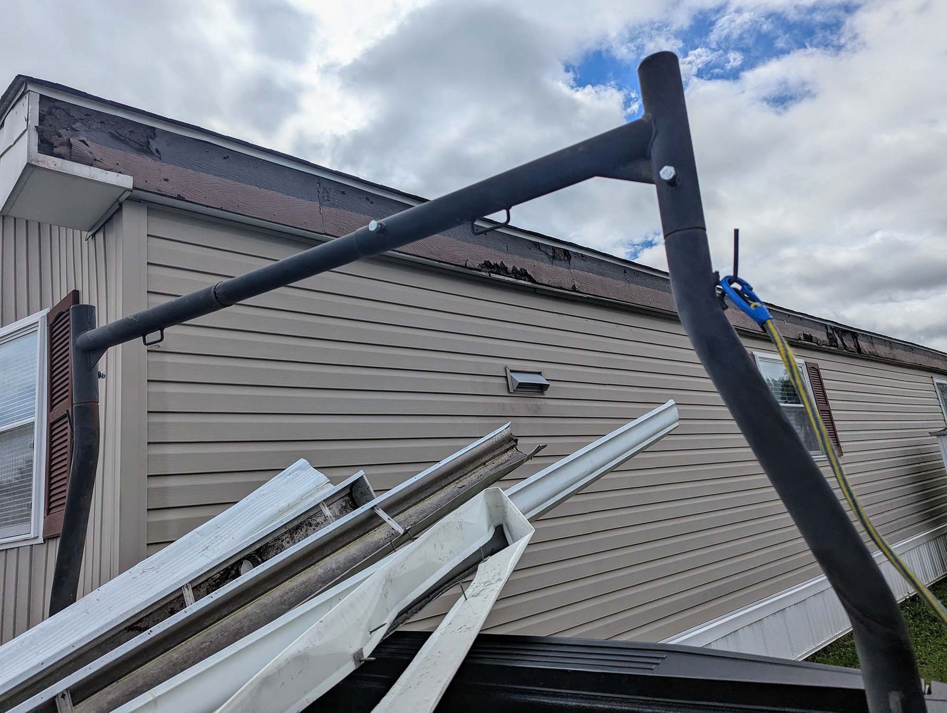 Black ladder rack on a truck bed, holding gutters, next to a beige house with a cloudy sky.