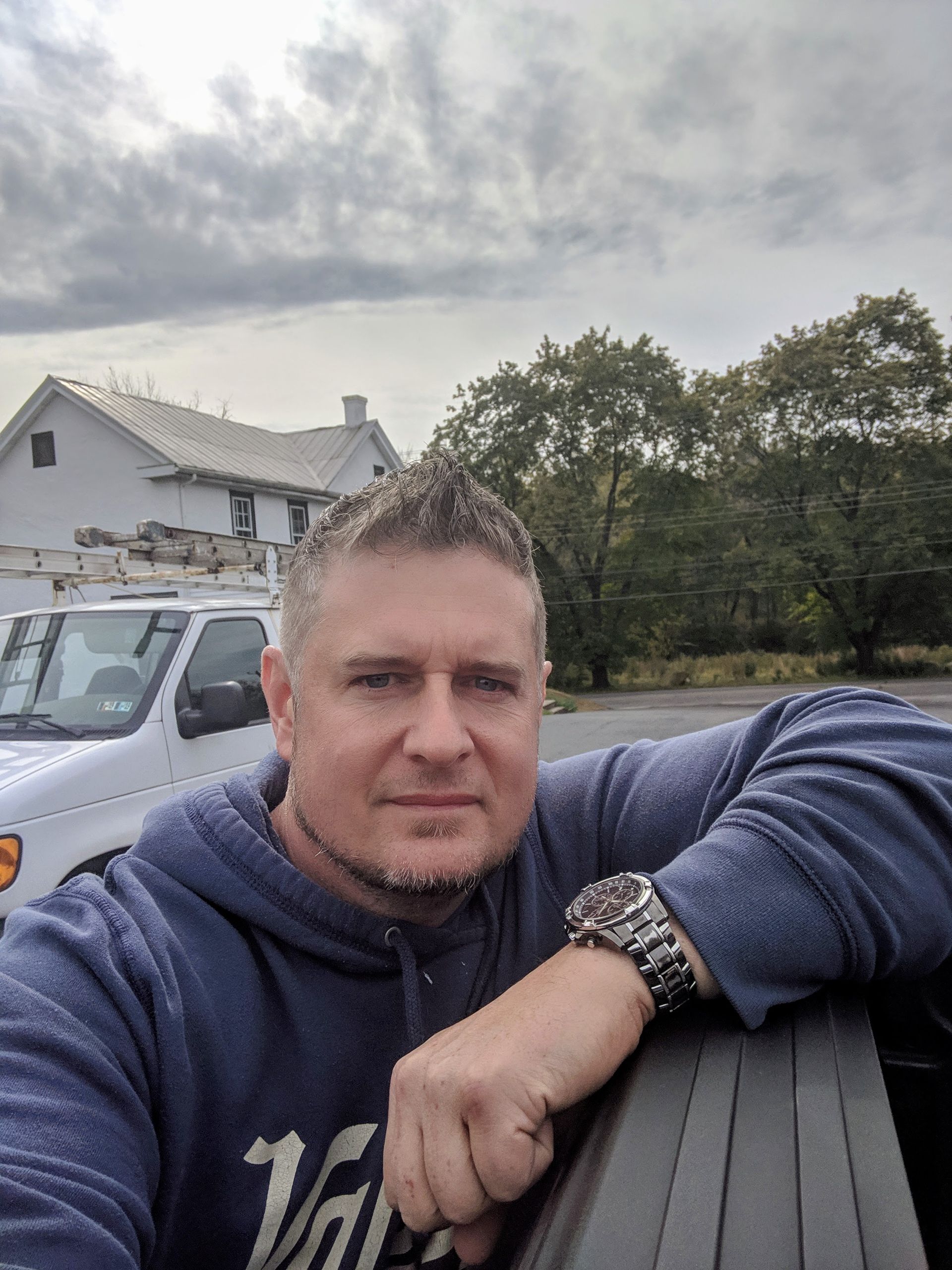 Man in blue hoodie leans against a truck, cloudy sky and building in the background.