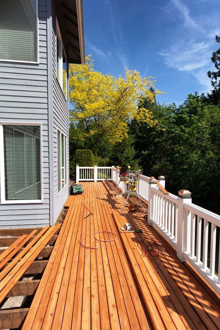 Wooden deck under construction next to a gray house, white railing, sunny with a yellow-leaved tree.