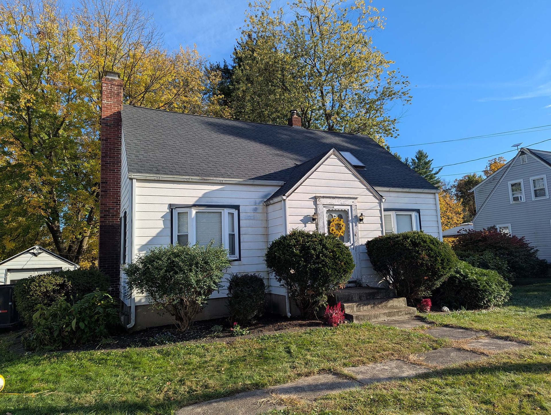 White house with dark roof, small front yard, bushes, chimney, sunny day.