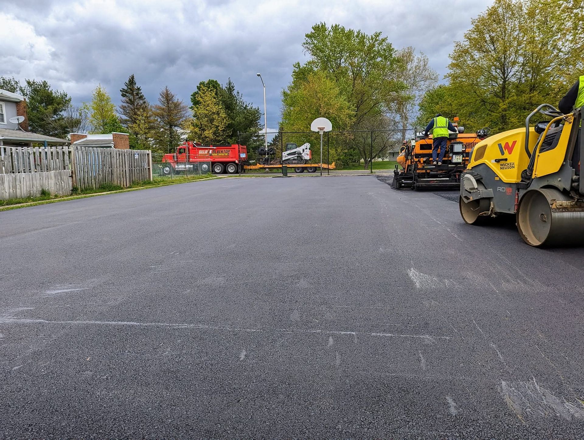 Asphalt paving of a basketball court with a red truck and two yellow rollers under an overcast sky.
