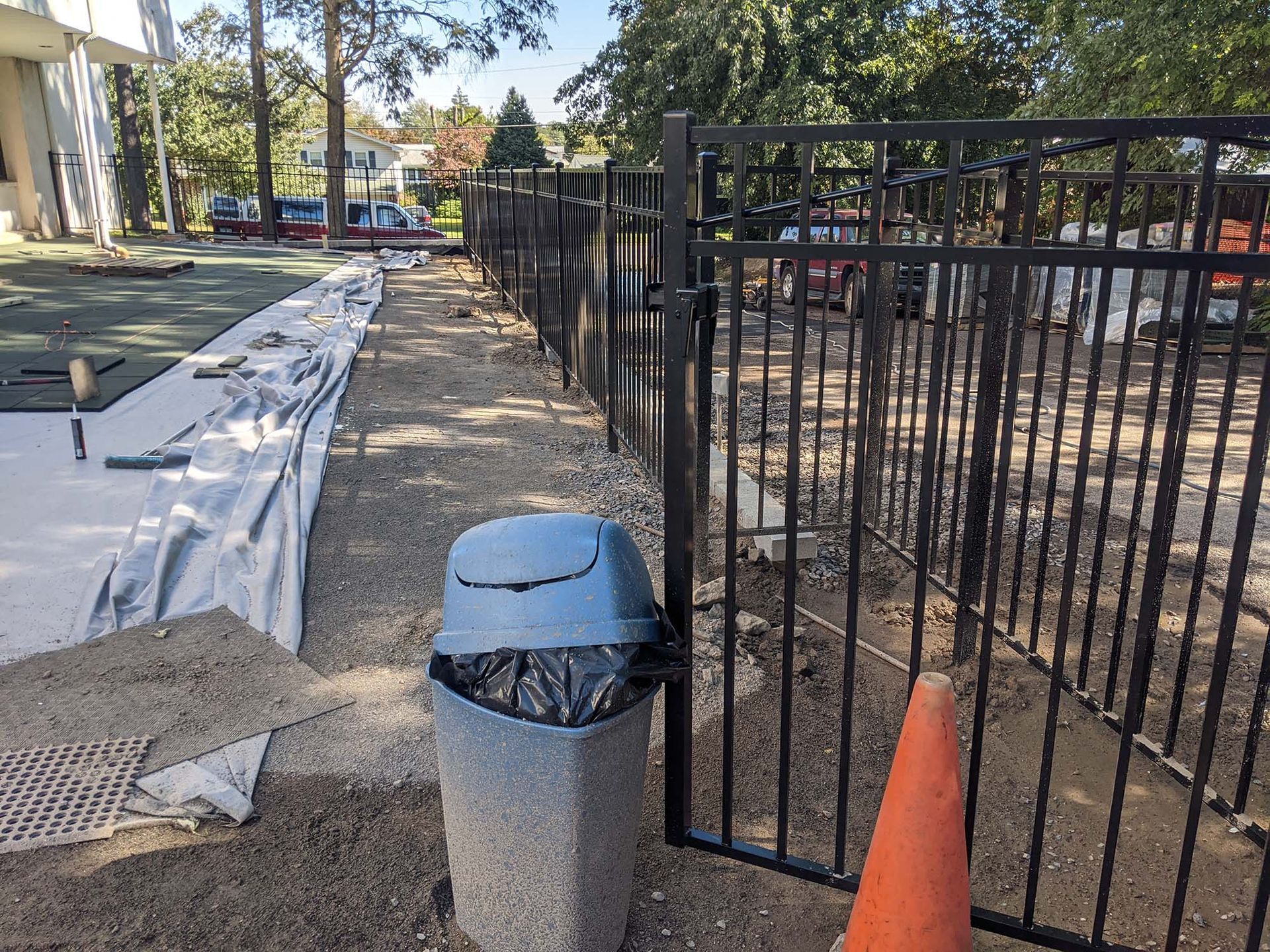 Black metal fence and gate beside a gravel pathway with a trash can and traffic cone.