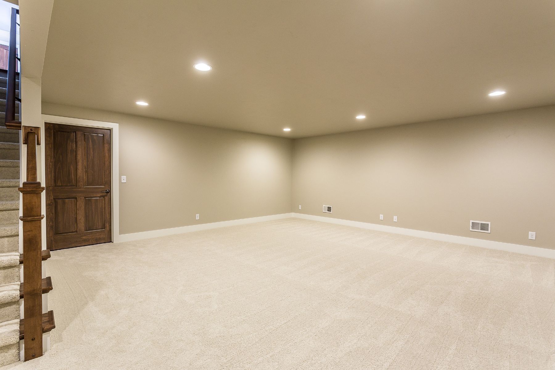 Empty beige-walled room with recessed lighting, brown door, and carpet. Staircase visible on the left.