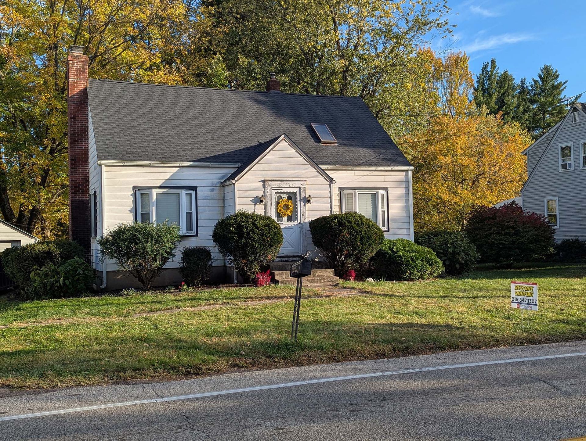 White house with black roof, small front yard, bushes, and a brick chimney. Autumn foliage in the background.