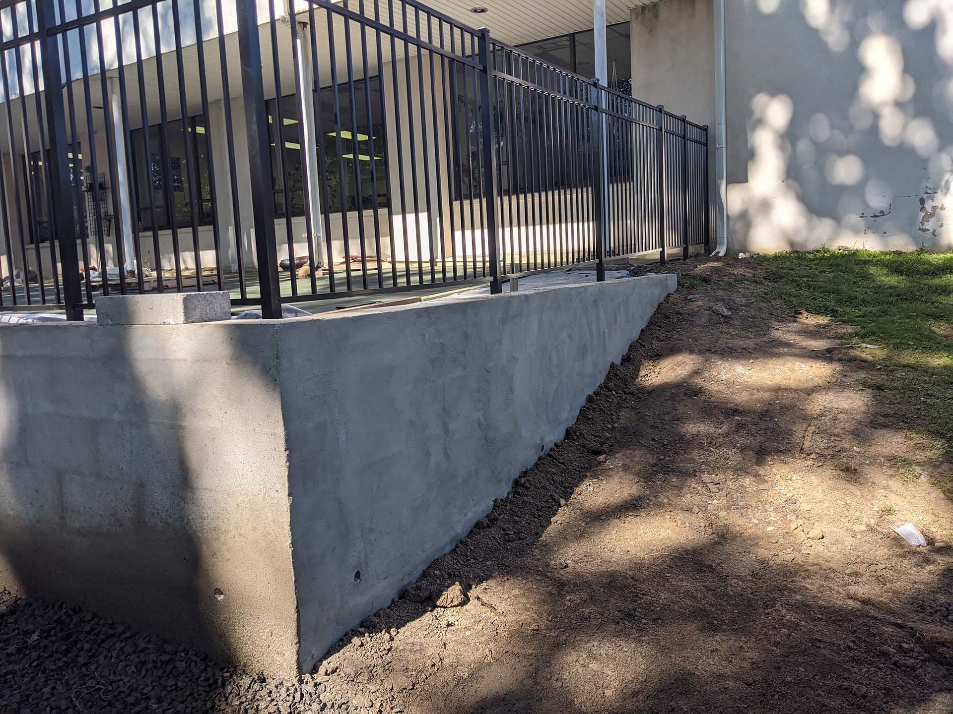 A concrete retaining wall with a black metal fence, next to a grassy area and dirt ground.