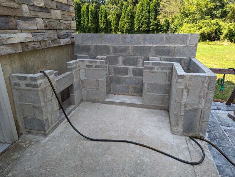 Outdoor brick structure under construction with a concrete floor, surrounded by a stone wall, and a garden backdrop.