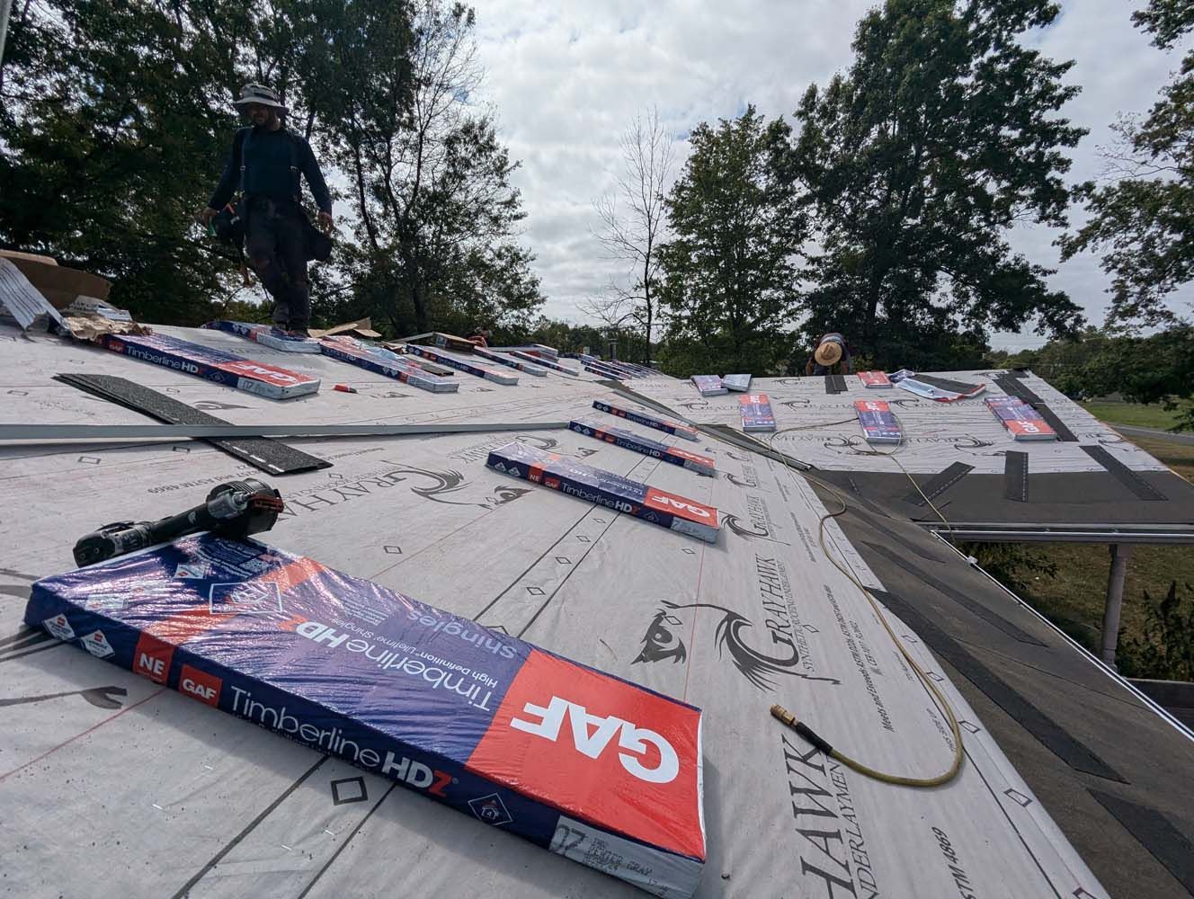 Roofer on a partially covered roof with bundled roofing materials. Tools and overcast sky are visible.