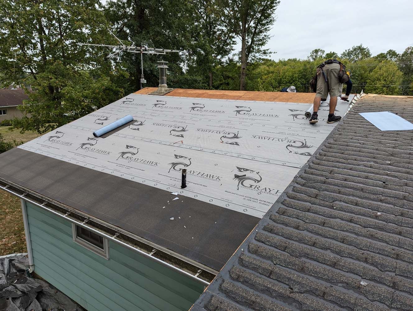 Person installing roofing material on a house roof. Gray shingles, green siding, overcast sky.