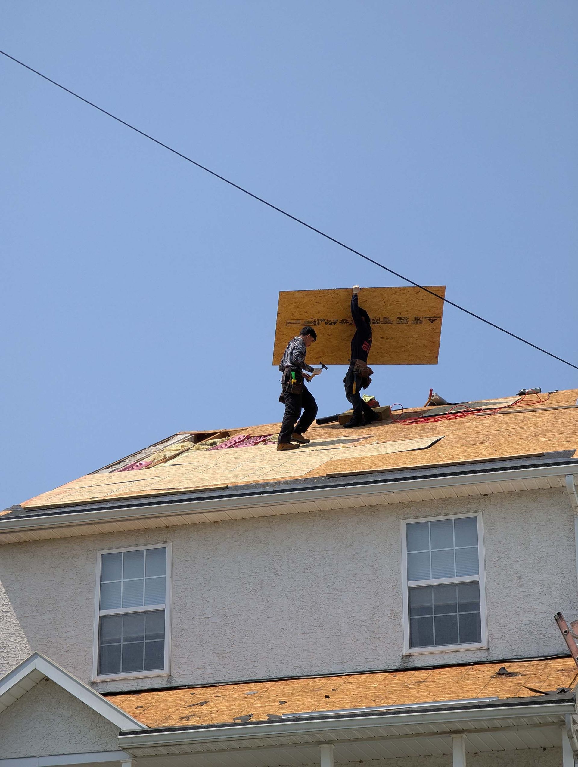 Two roofers carrying plywood on a rooftop under a bright blue sky.