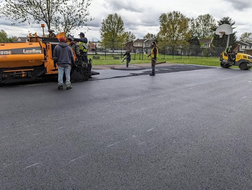 Road paving: Workers and machinery laying asphalt on a paved surface, cloudy sky overhead.