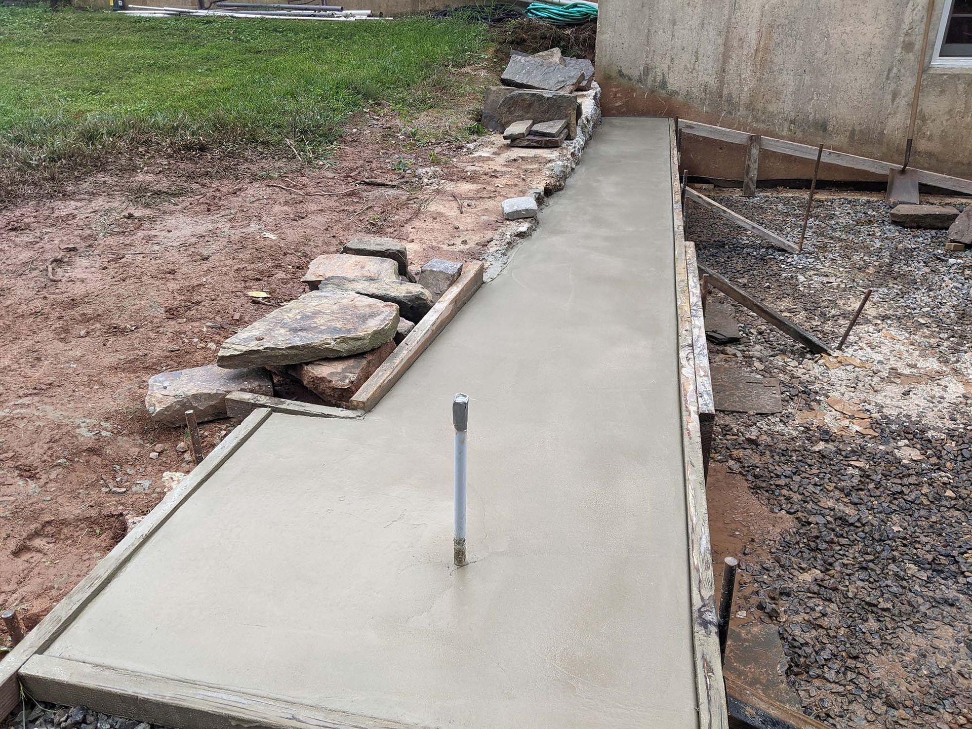 Freshly poured concrete walkway next to a building. Stone border and unfinished landscaping in the background.