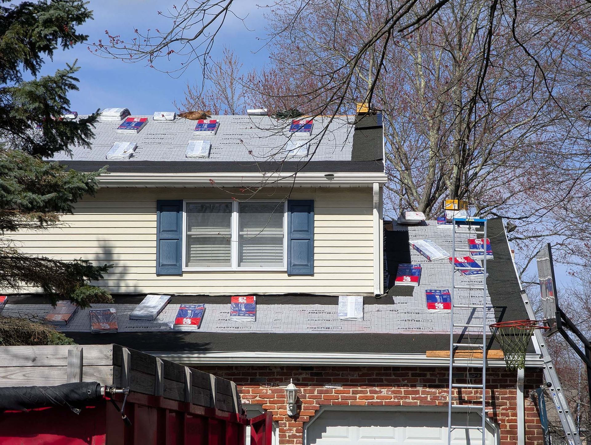 House roof partially re-shingled. Sections covered with new shingles and safety flags. Ladder beside roof.