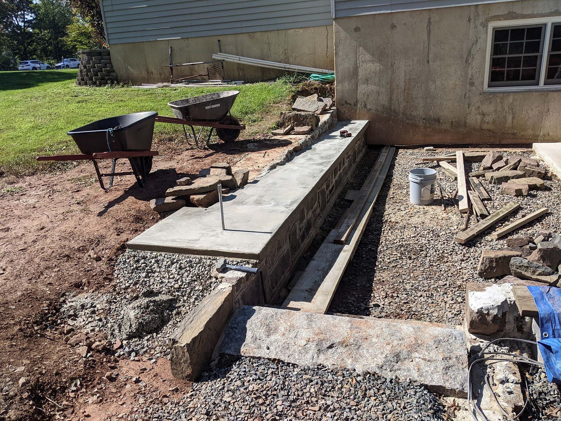 Construction site: a concrete retaining wall under construction next to a building. Wheelbarrows, tools, and gravel visible.