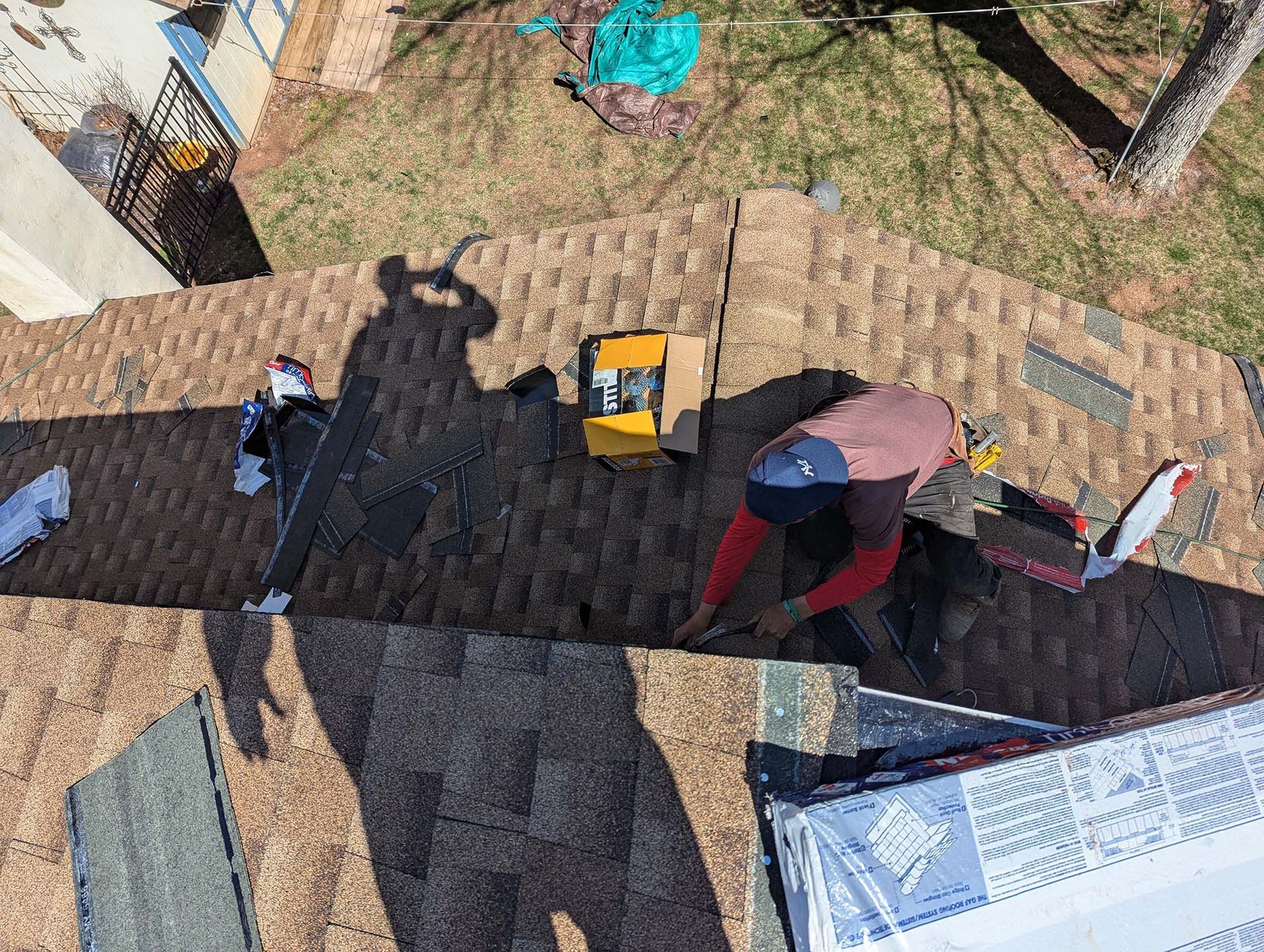 Roofer on a brown shingled roof, working with tools and materials. Sunny day, overhead view.