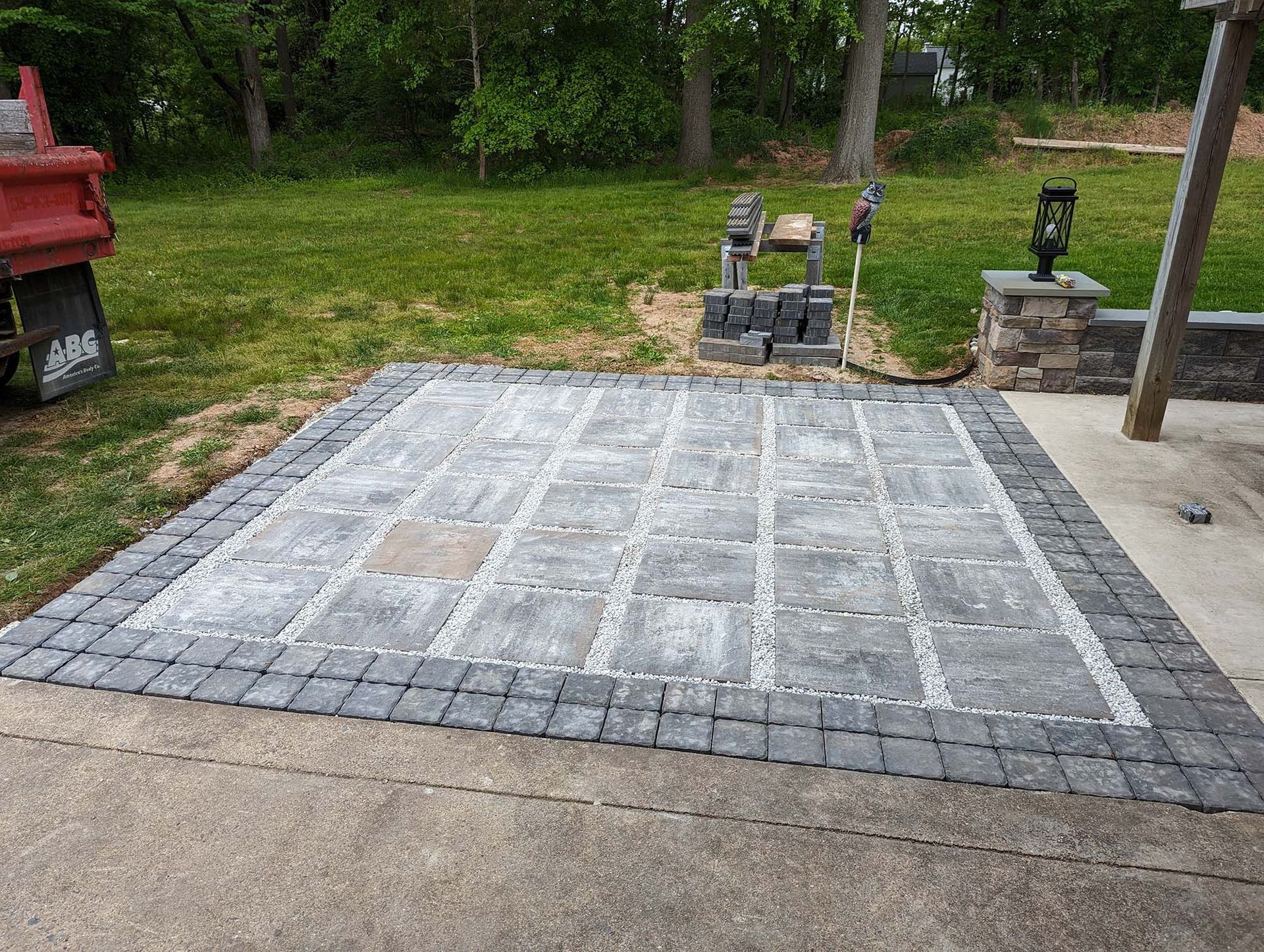 Newly paved patio with gray and black pavers, bordered by a concrete slab. A red truck is visible to the left.