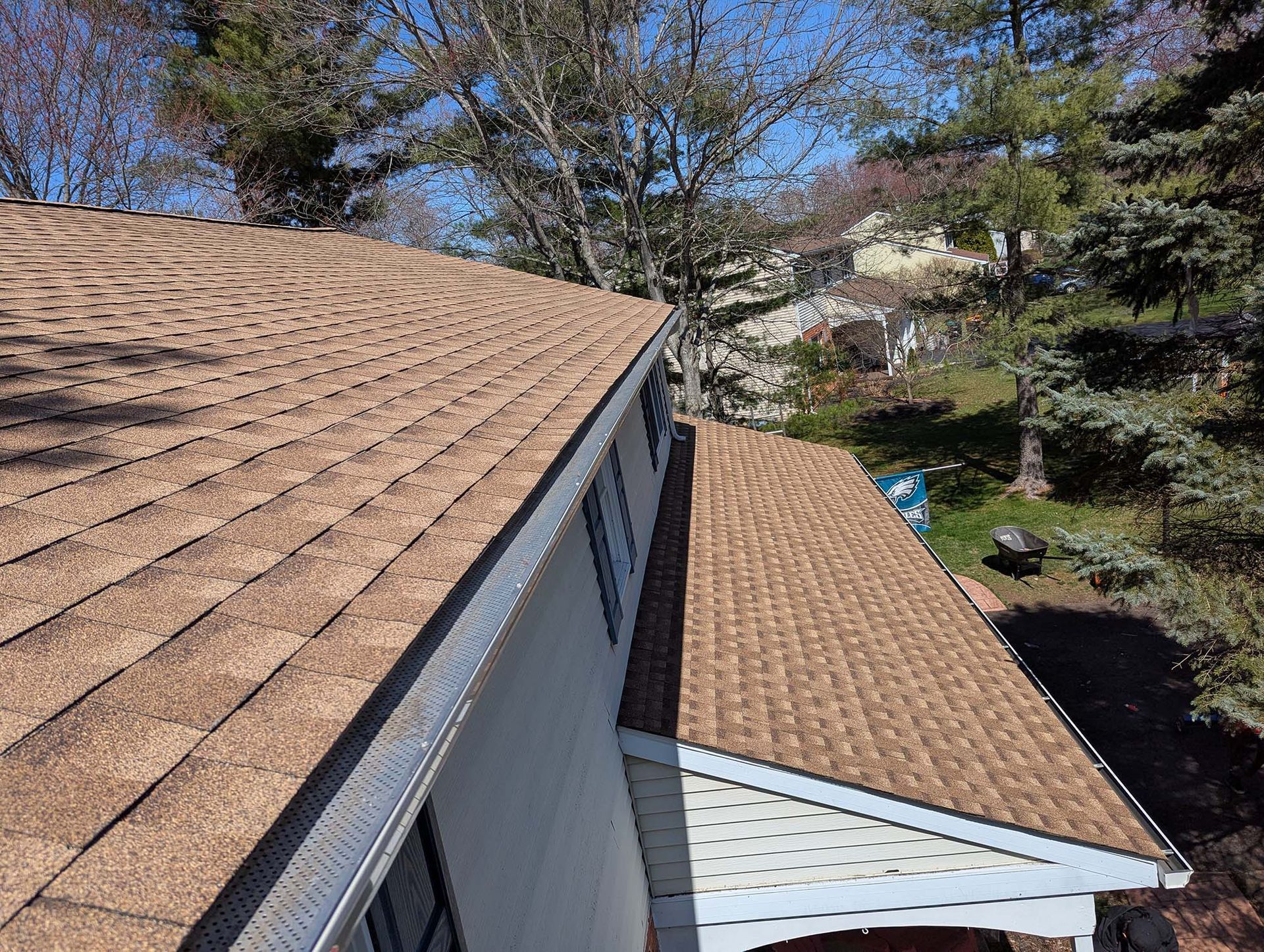 Brown asphalt shingle roof on a house, angled view, with trees and a blue sky in the background.