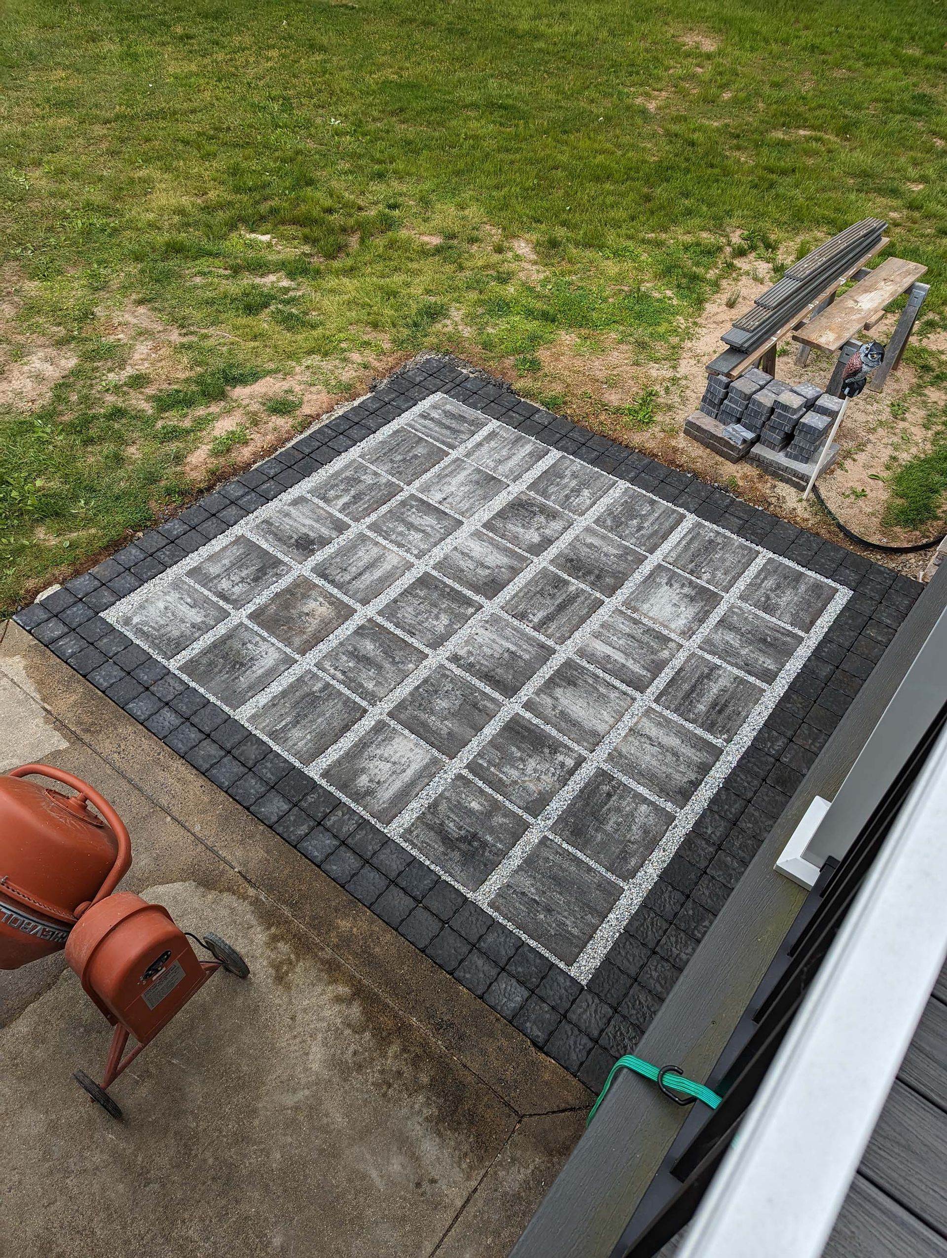 Square patio paved with gray and black stones, bordered by black bricks.  Nearby, more bricks are stacked.