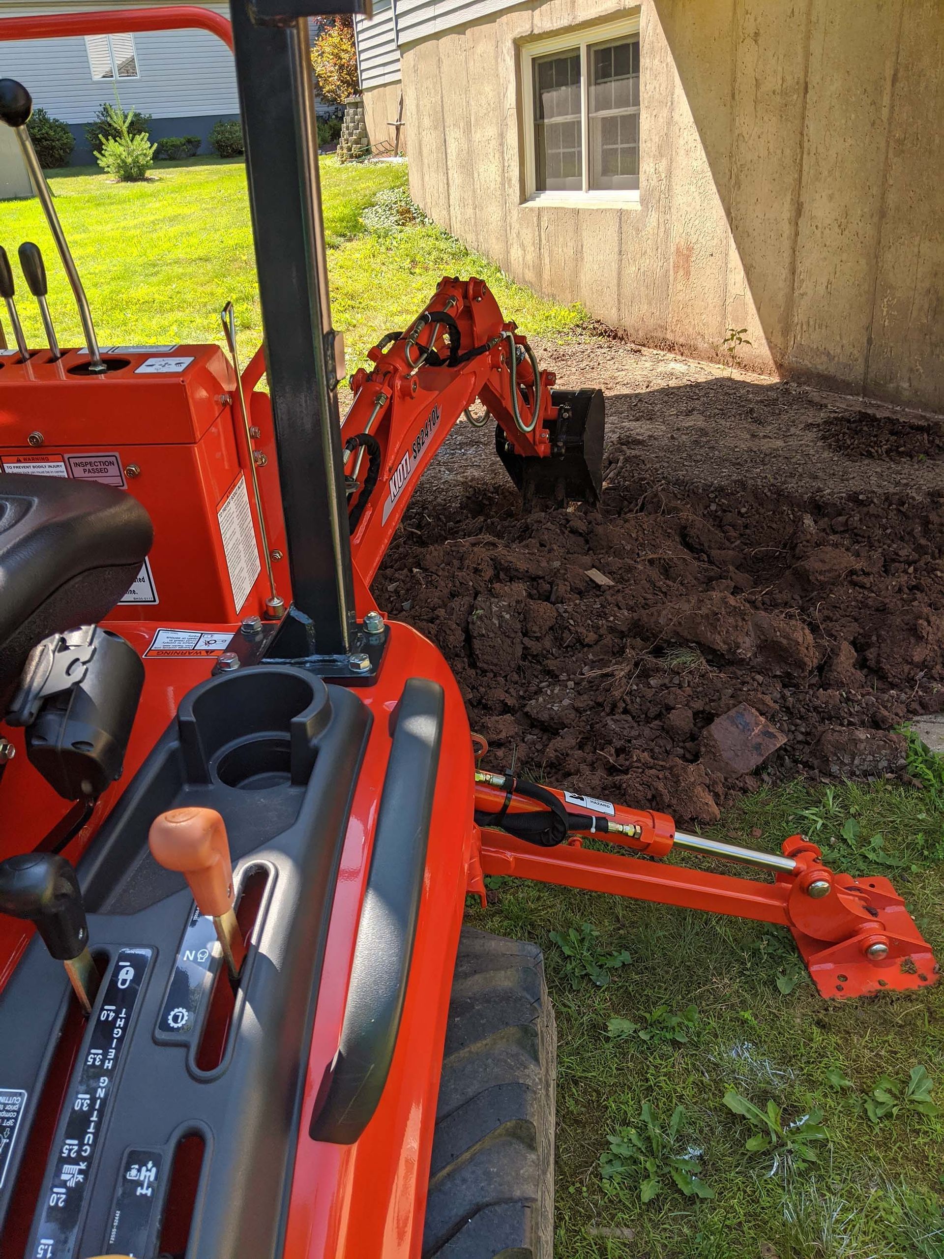 Orange tractor with backhoe digging in dark soil next to a building.