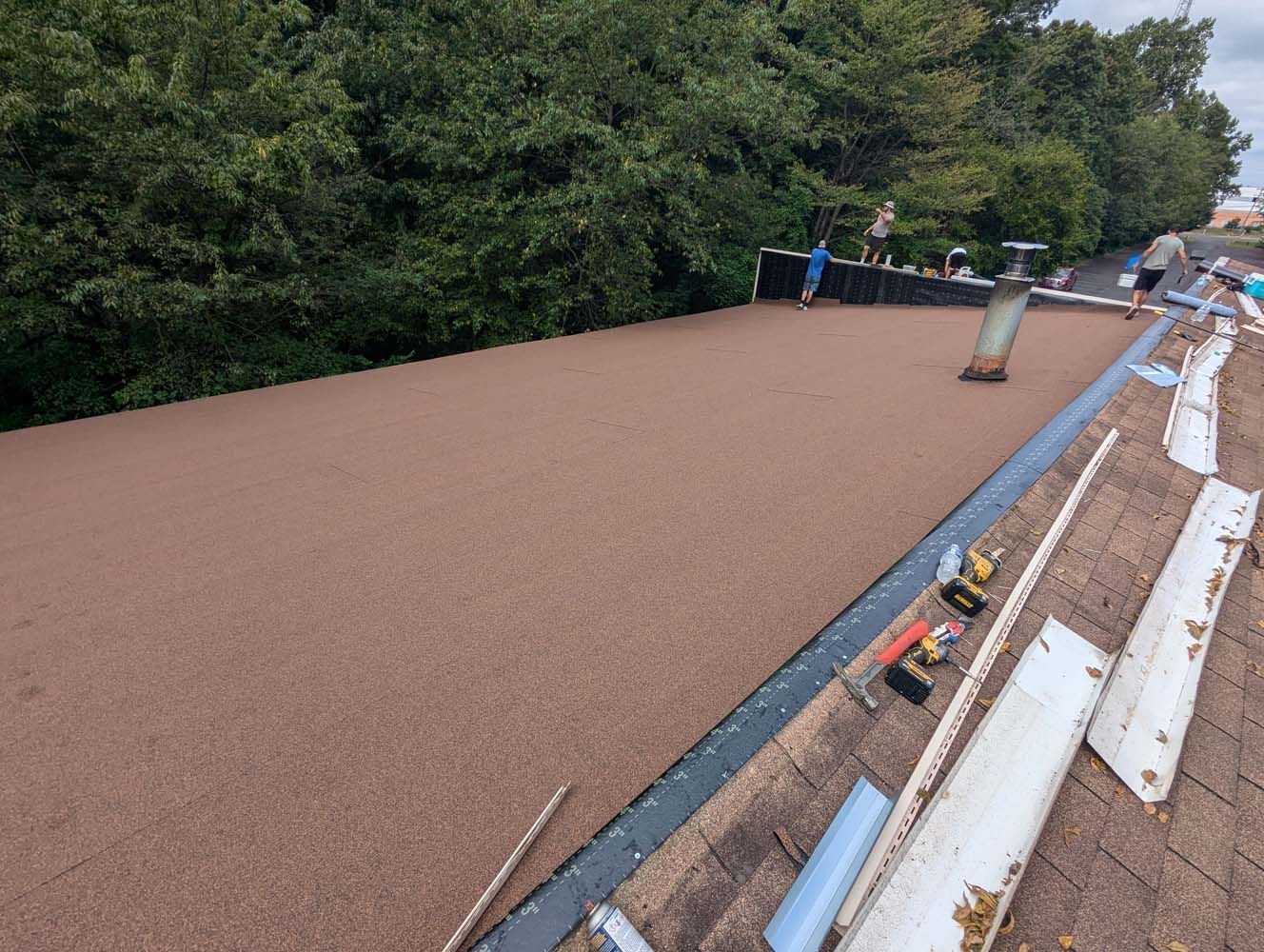 Brown gravel-covered flat roof with workers. Trees in the background, tools and materials visible on the roof.