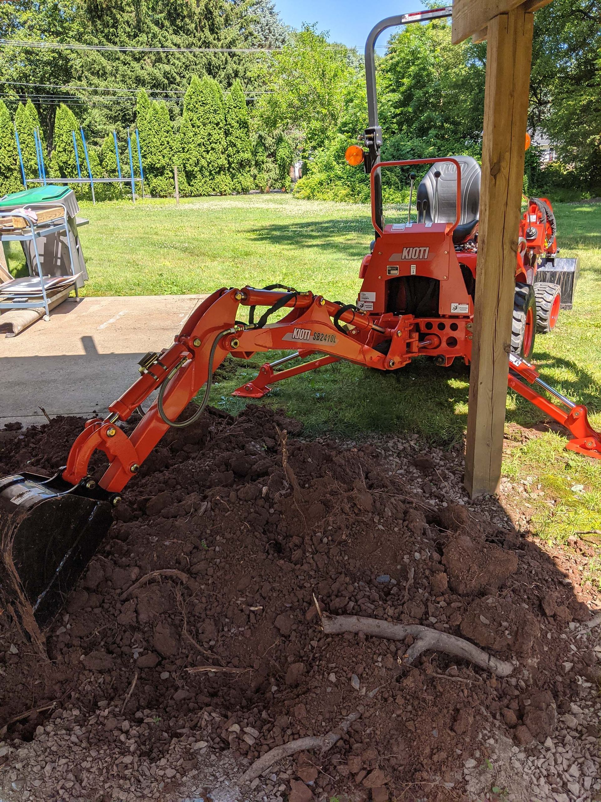 Orange tractor with a backhoe digging in a pile of dirt next to a wooden post.