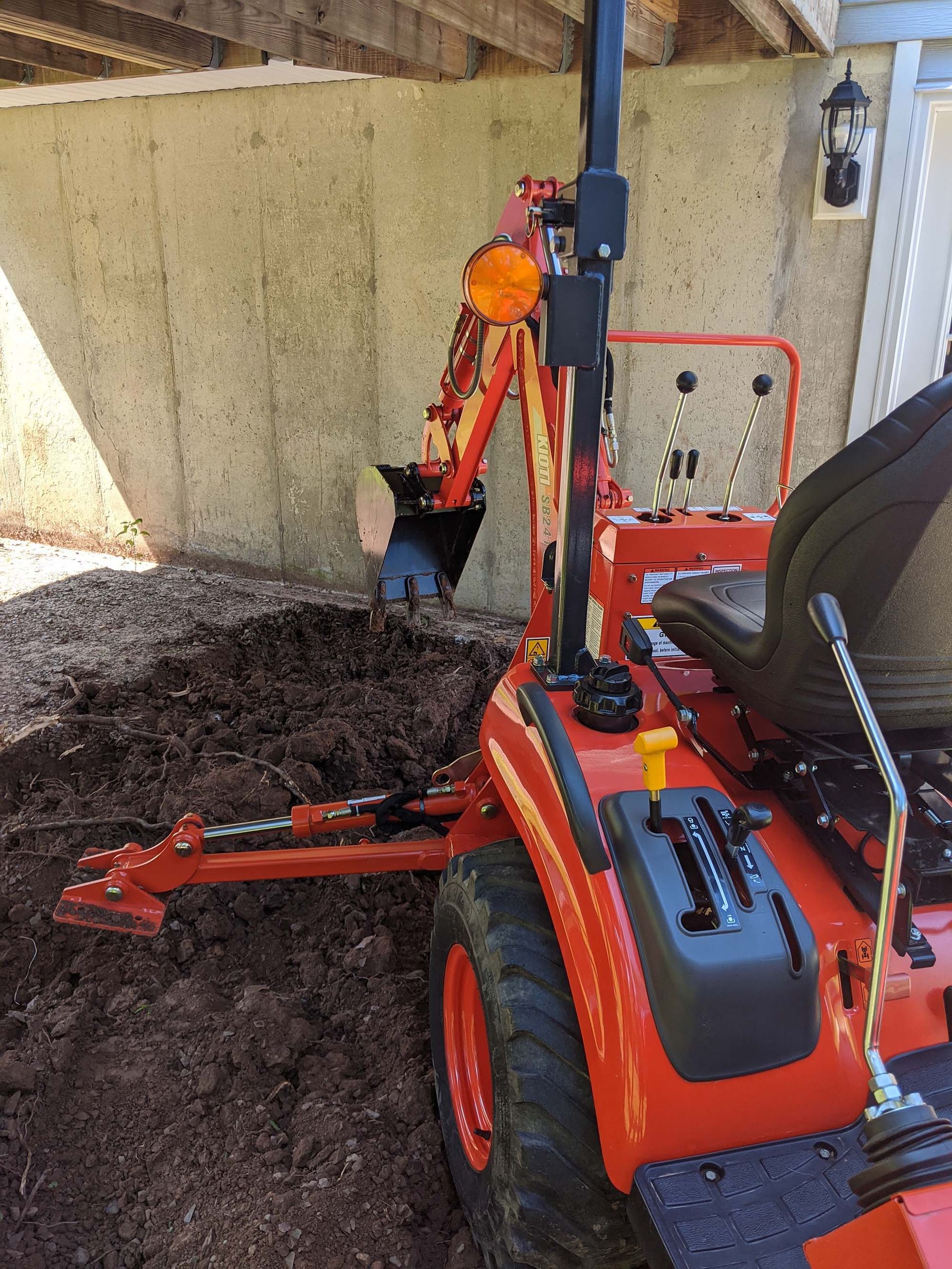 Orange tractor with backhoe digging in dark soil under a concrete structure.