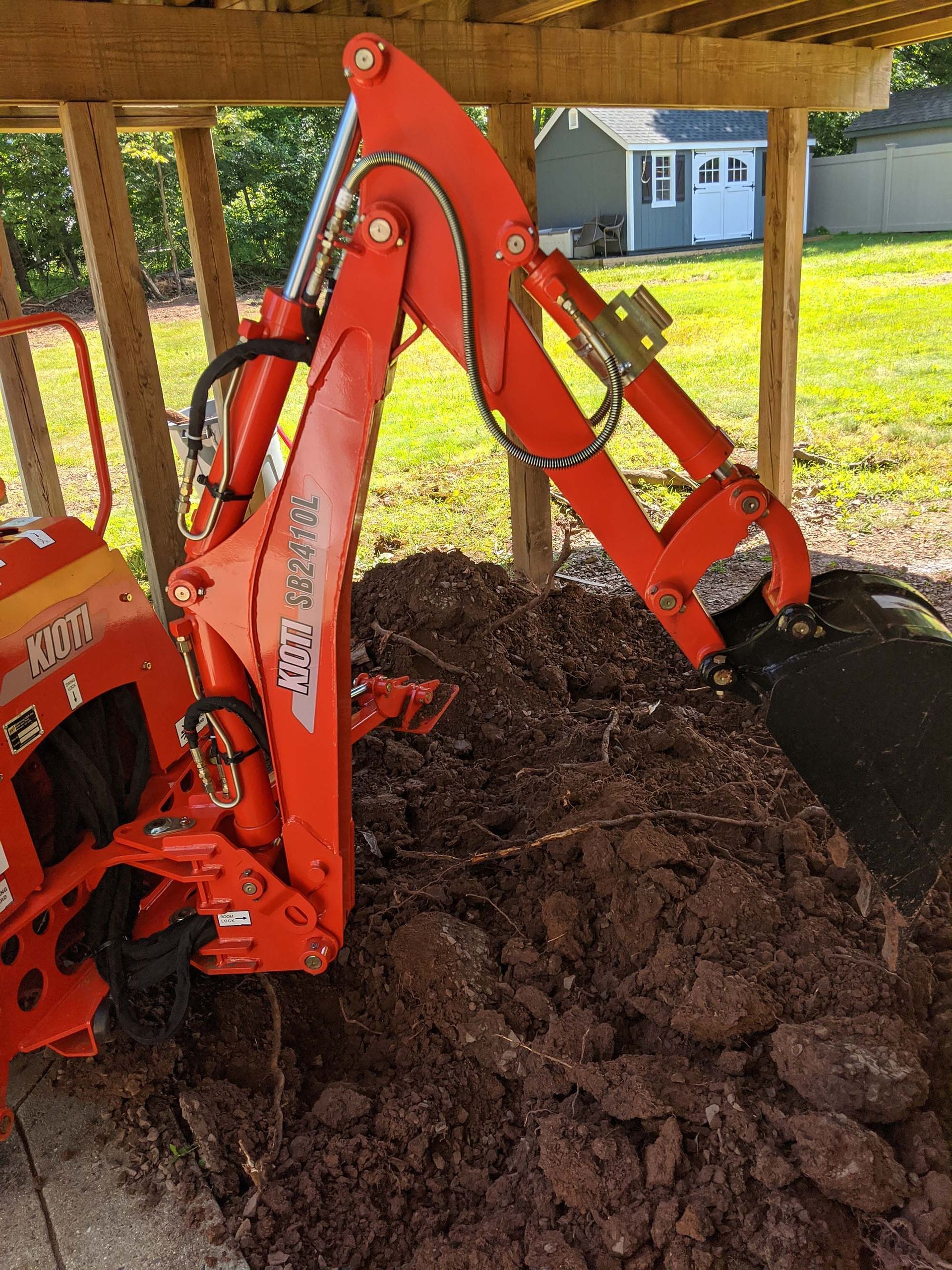 Orange backhoe digging in dark brown soil under a wooden structure.