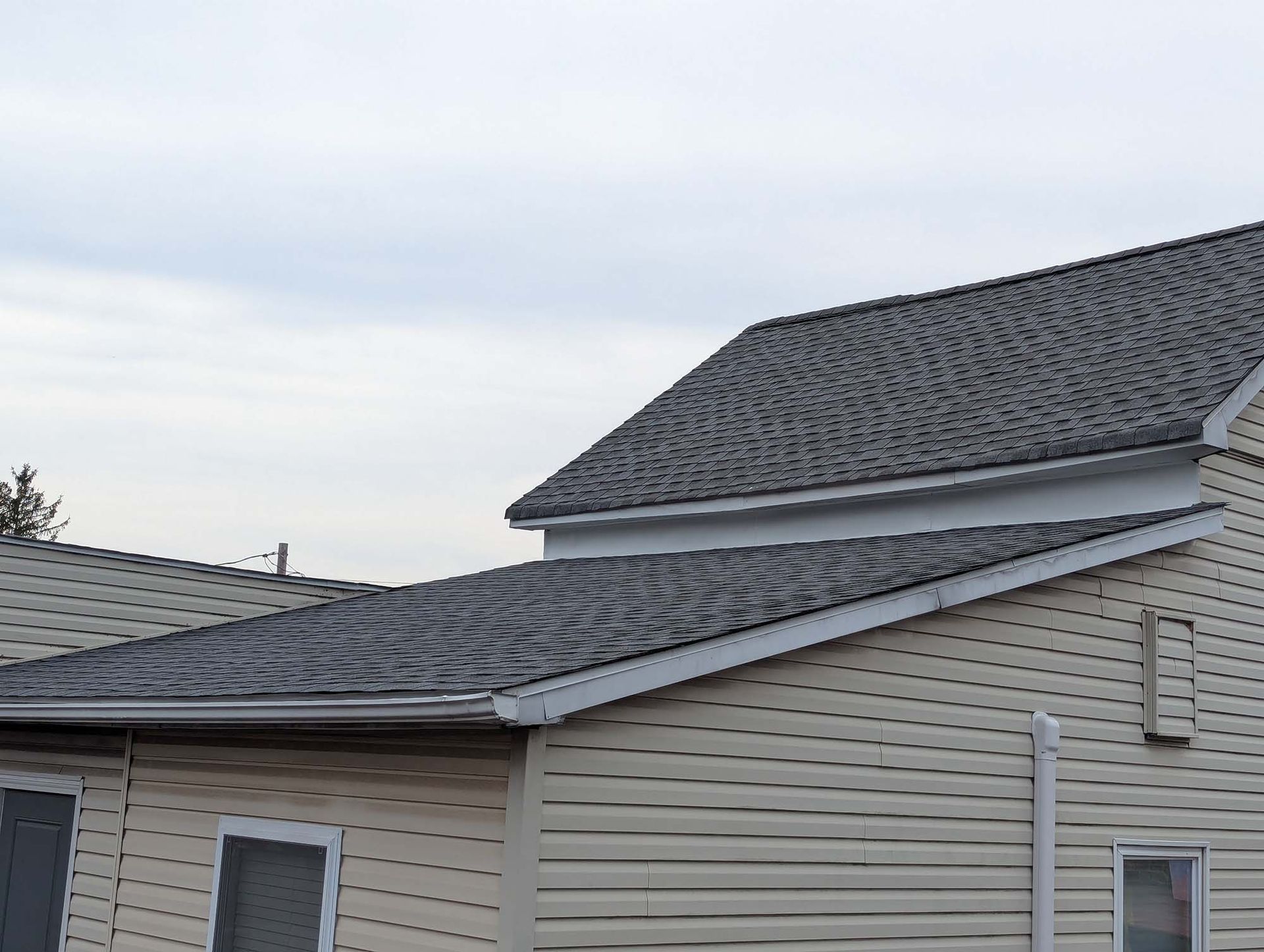 House with gray shingled roof and tan siding under a cloudy sky.