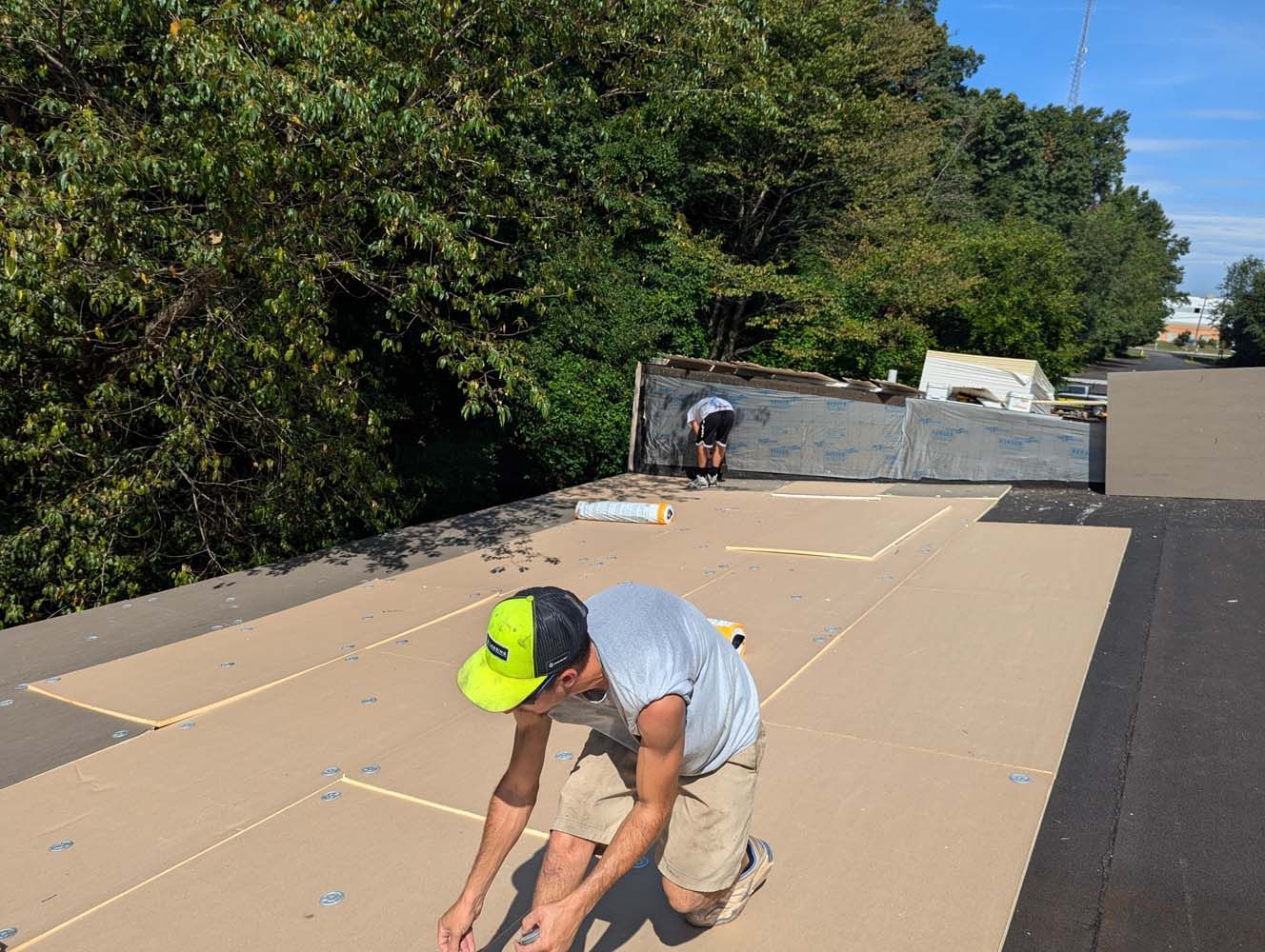 Two workers on a flat roof; one kneels, the other works near the edge. Brown paper and tools are visible.
