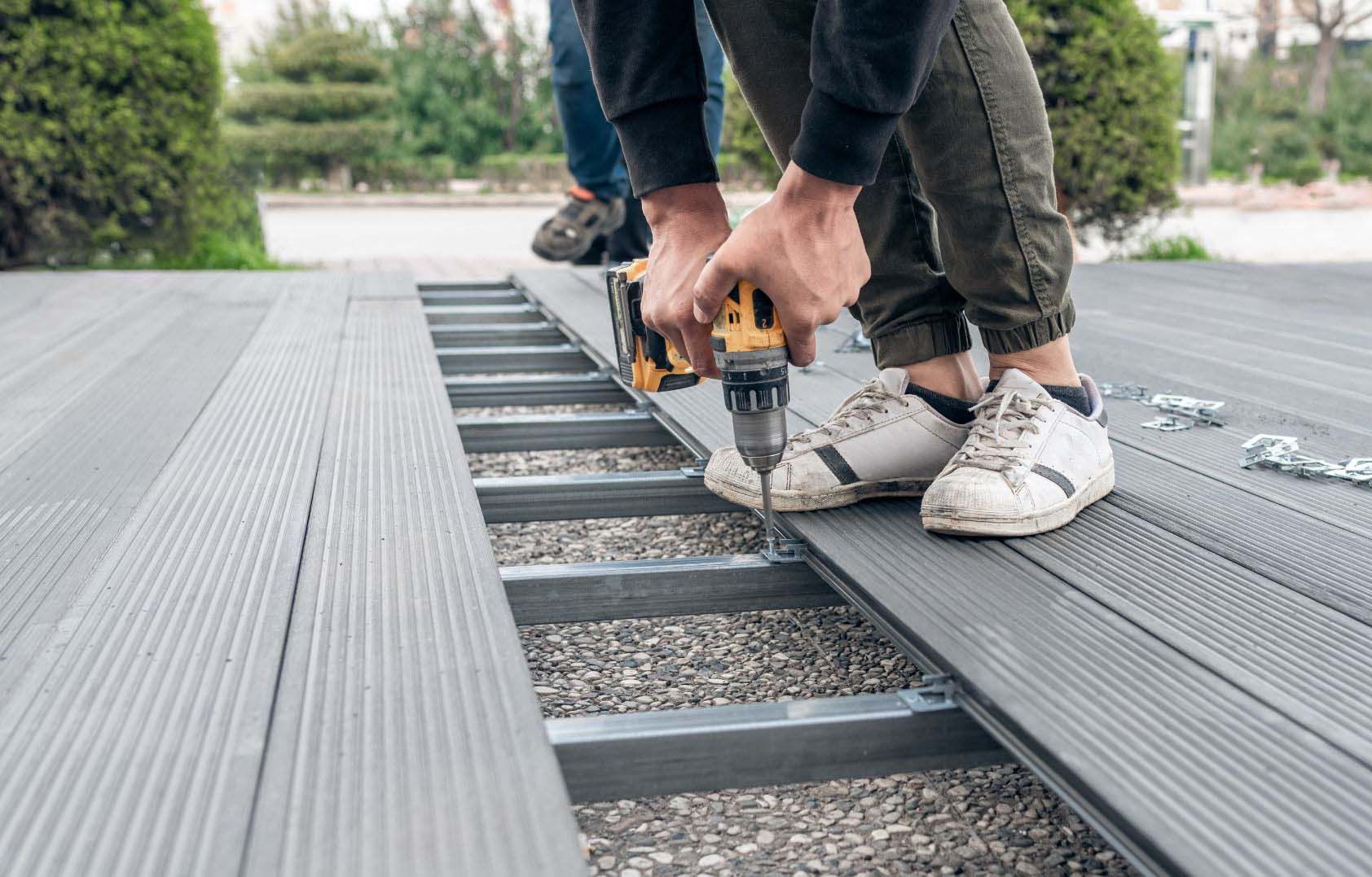 Person using a drill to install a gray deck board on a metal frame, outdoors.