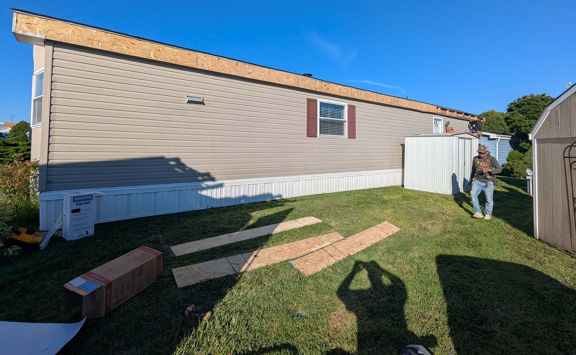 Man working on a mobile home's roof edge. Boards and cardboard on the grass. Sunny day.