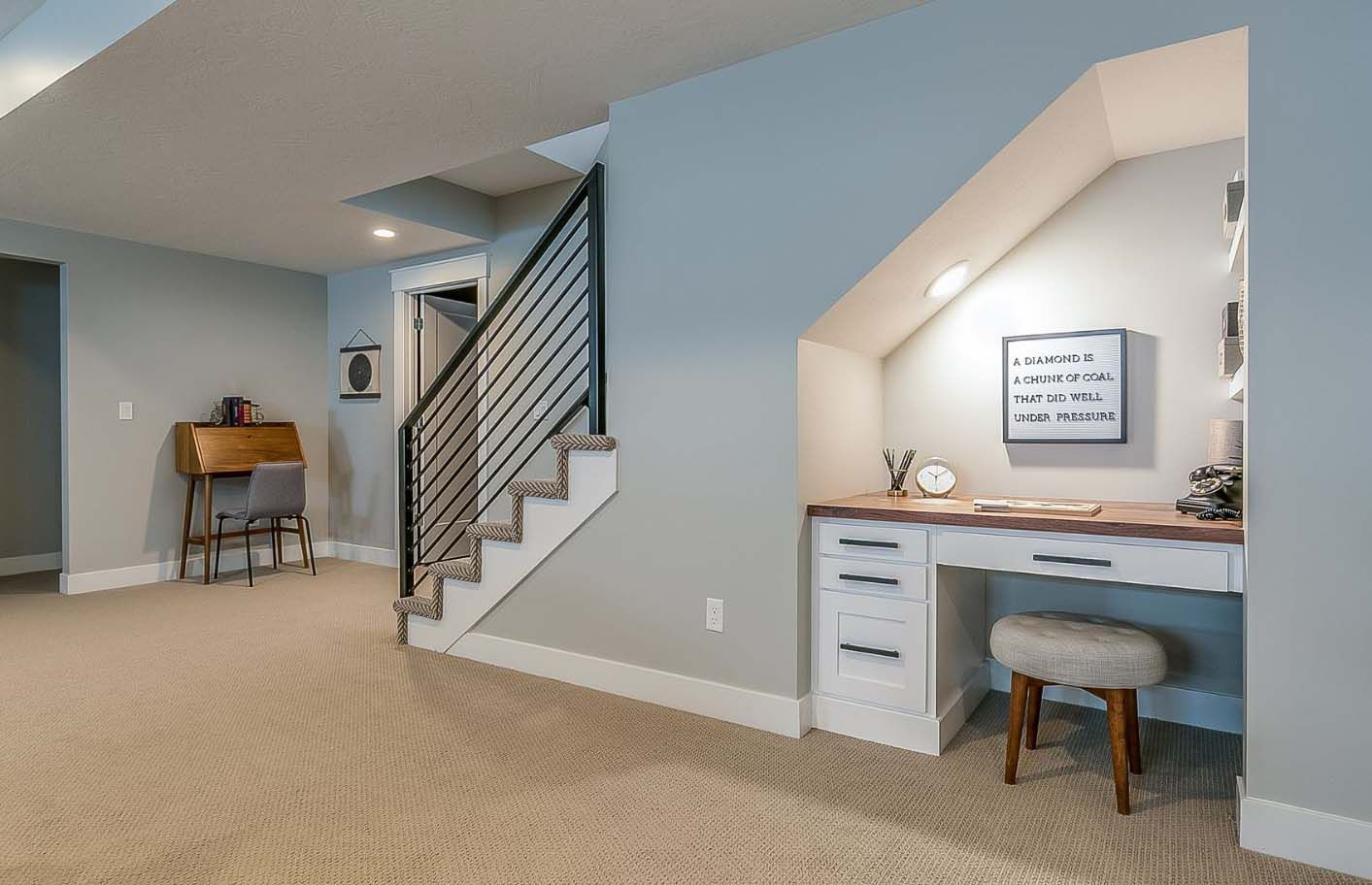 Basement home office with desk built into the wall under the stairs; neutral colors, carpeted floor.
