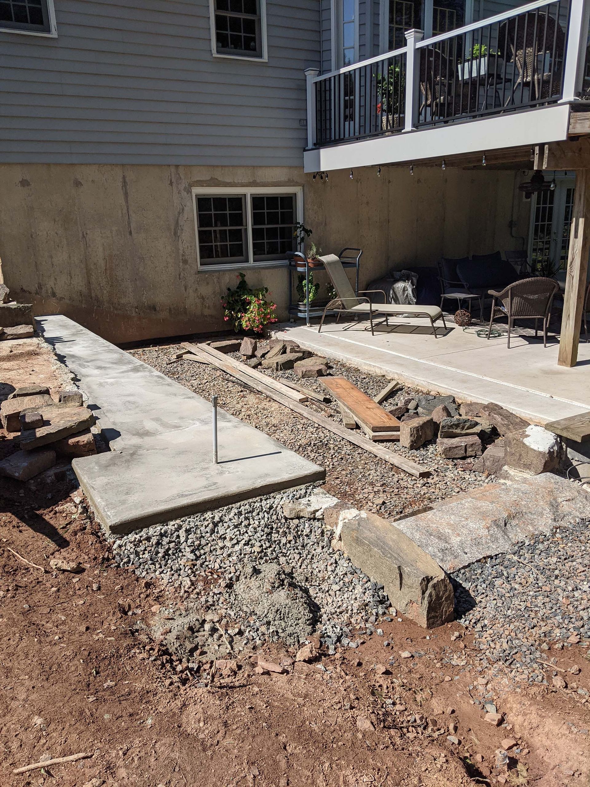 Construction site: excavation for a patio or walkway next to a house with a deck. Gravel and concrete visible.