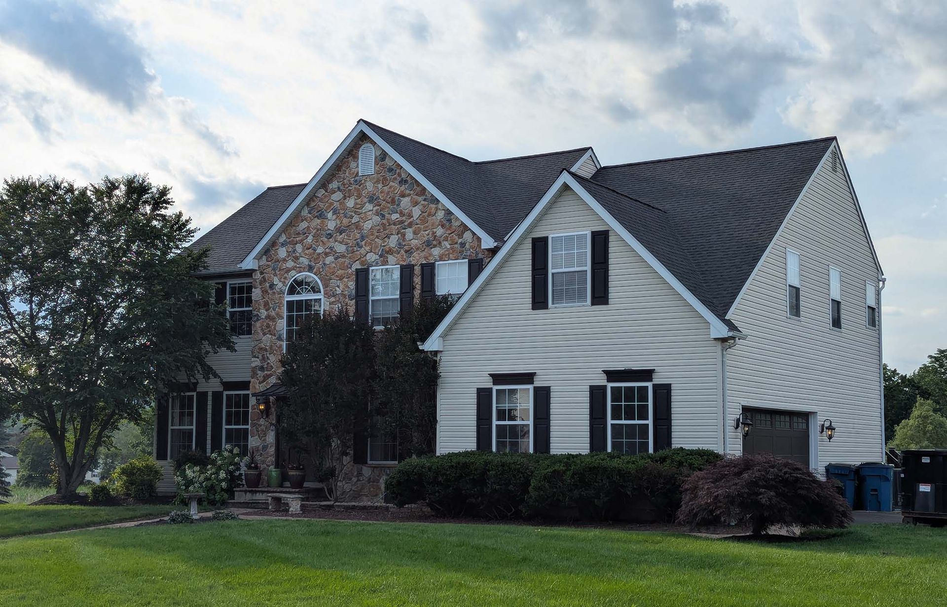 Two-story house with stone and white siding, dark shutters, and a dark roof. Green lawn and cloudy sky.