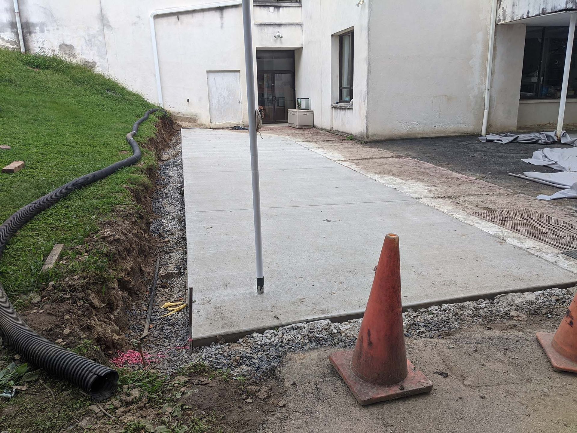 New concrete pathway next to building, with an orange traffic cone in the foreground.