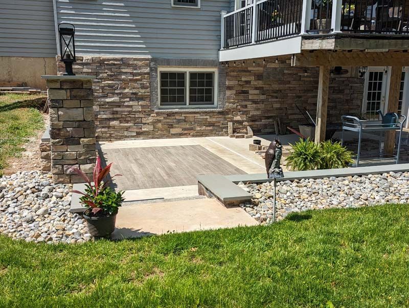 Stone patio and walkway by a house with a stone veneer, surrounded by landscaping, grass, and rocks.