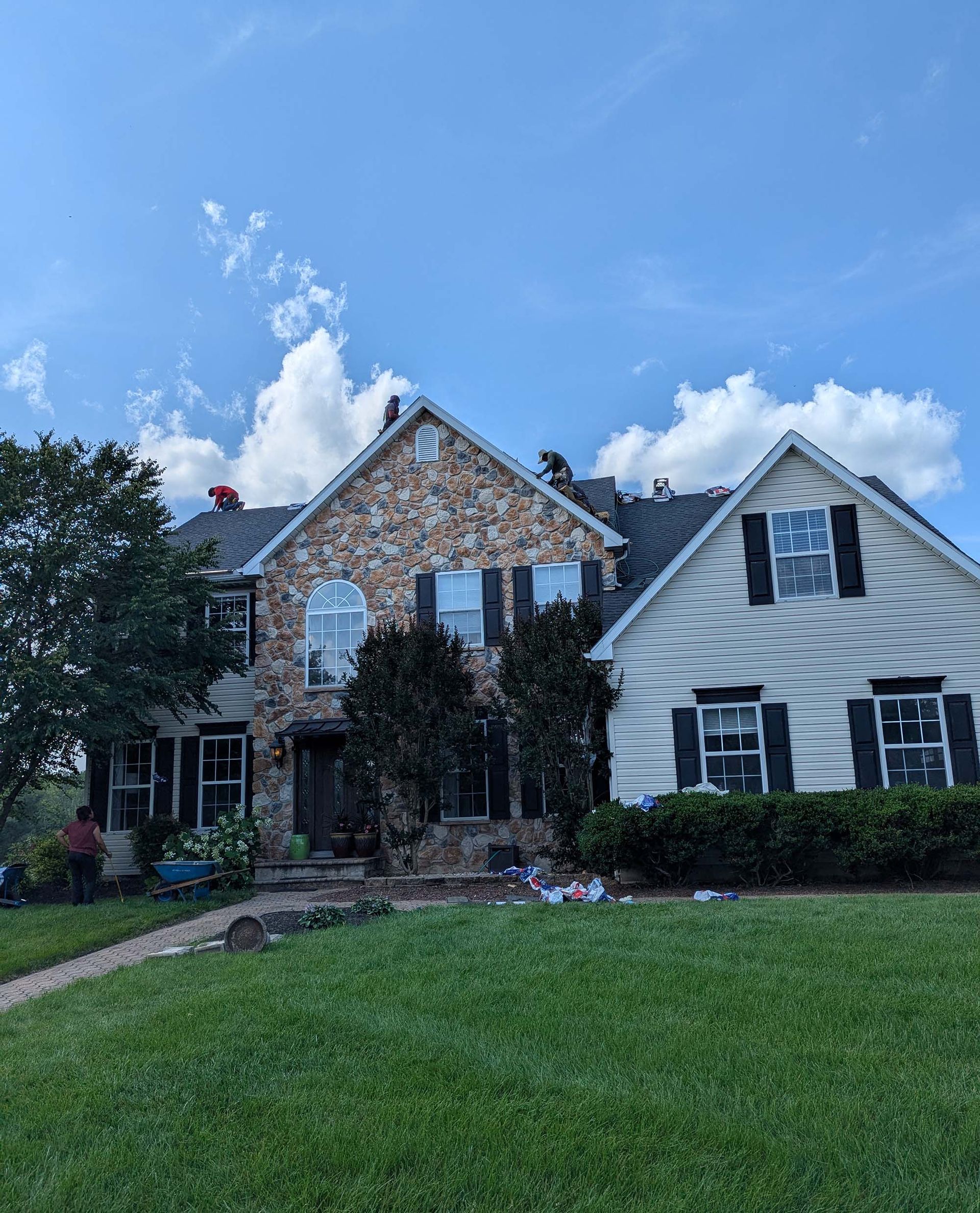 Two-story house with stone and siding facade, workers on roof, blue sky and green lawn.