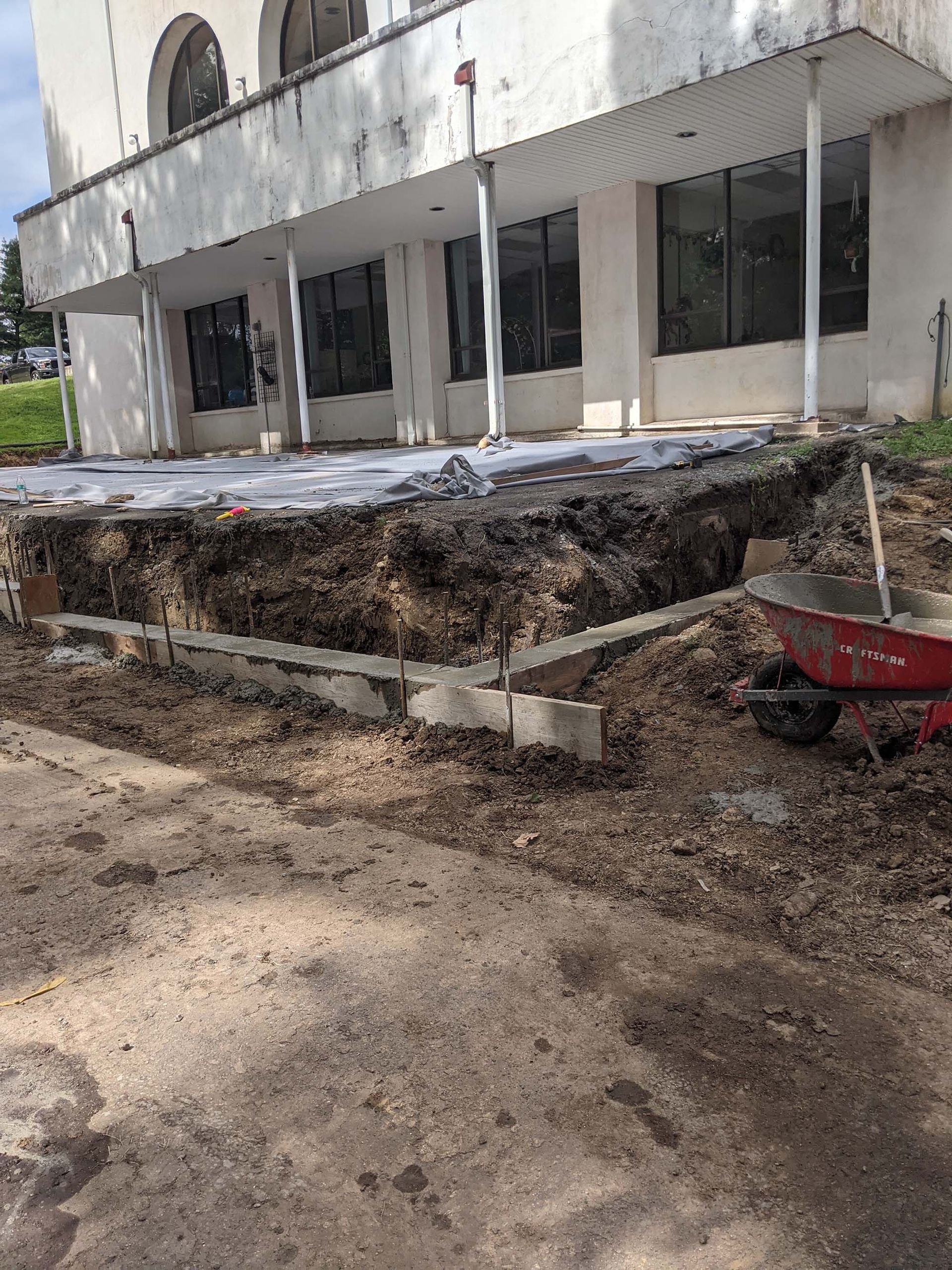 Construction site: foundation work in progress near a white building with large windows and a covered porch; dirt, concrete, and a wheelbarrow visible.
