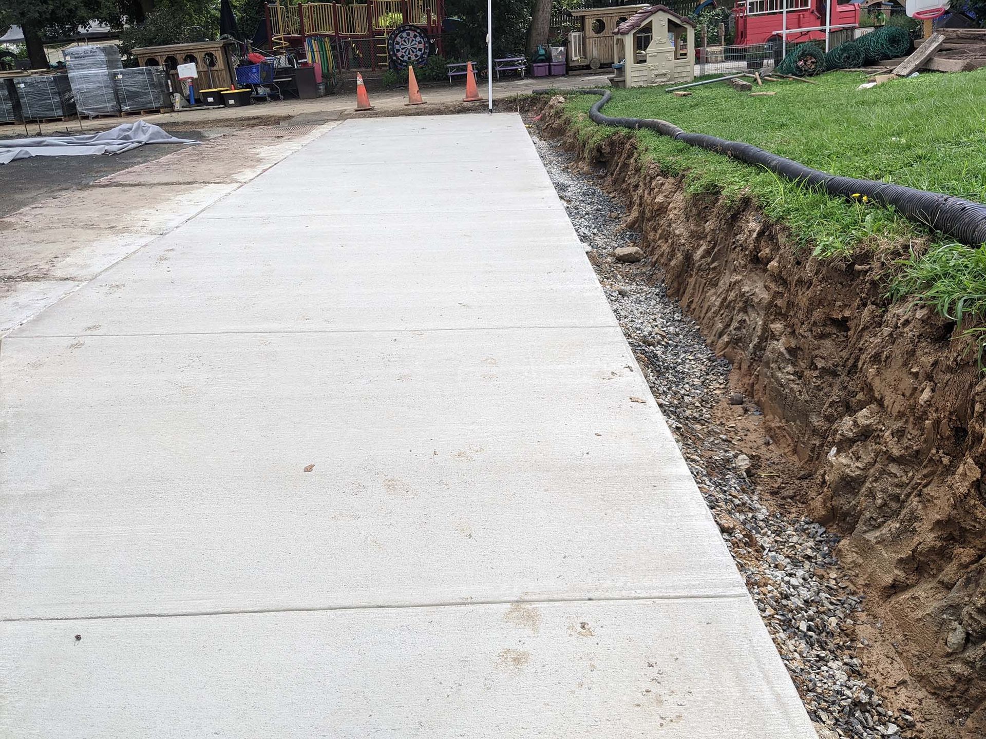 Concrete walkway next to excavated area with gravel. Black hose visible, green grass in background.