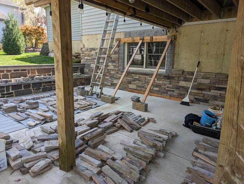 Construction site: Stone veneer installation on a building's exterior. Stone piles, ladder, and support beams visible.