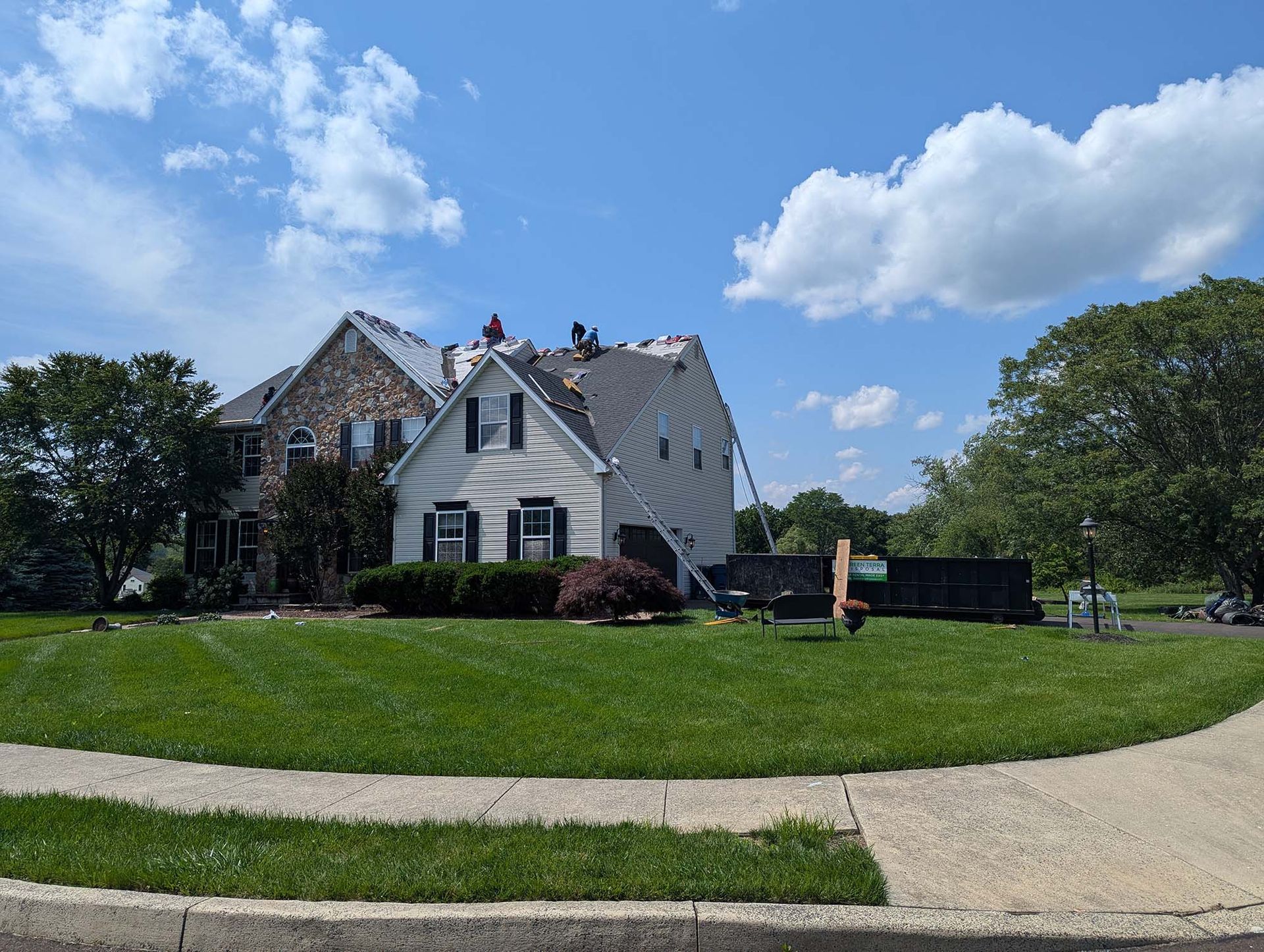 House with roofers replacing shingles on a sunny day. Green grass and blue sky.