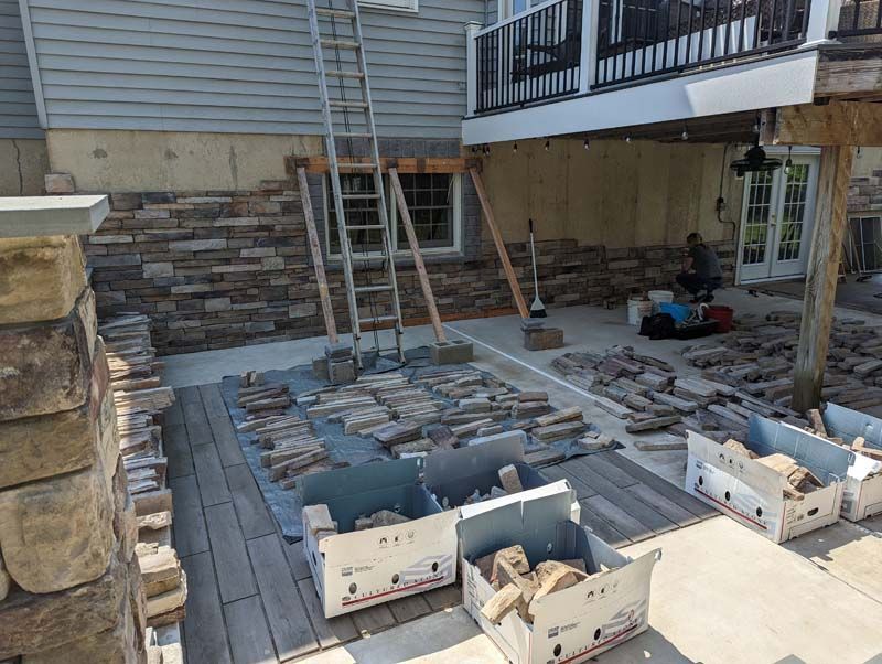 Stone siding being installed on a house exterior.  Ladder, tools, and boxes of stone are present.