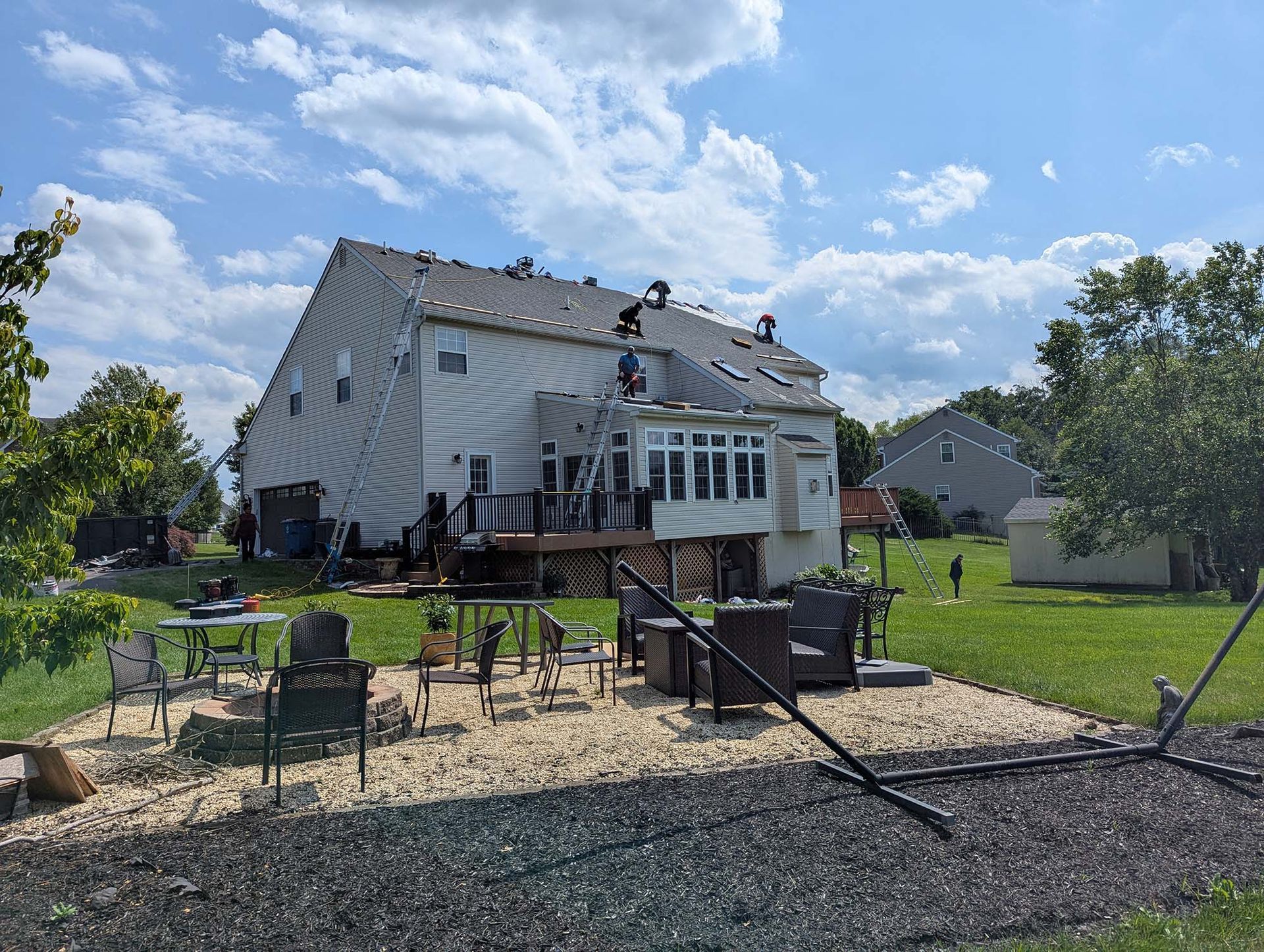 Backyard view of a two-story house with people on the roof, sunny day. Deck, patio furniture, and lawn in view.