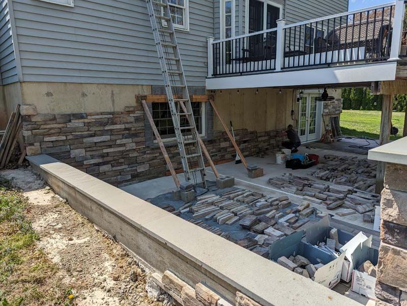 Construction of stone veneer on a house's exterior wall and patio. Ladder leaning against the wall. Materials and worker visible.