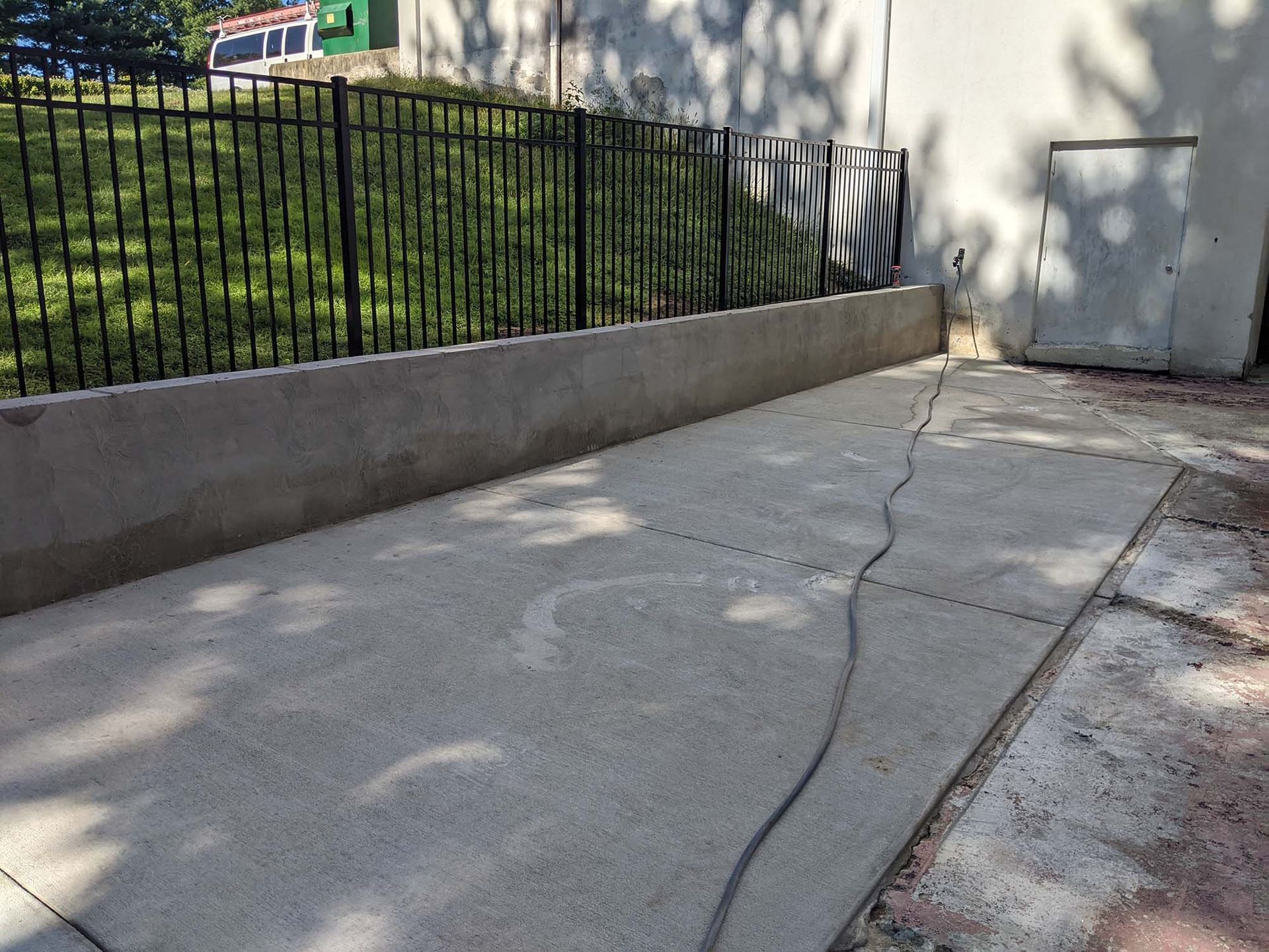 Concrete patio next to a concrete wall and a black metal fence, casting shadows on the surface.