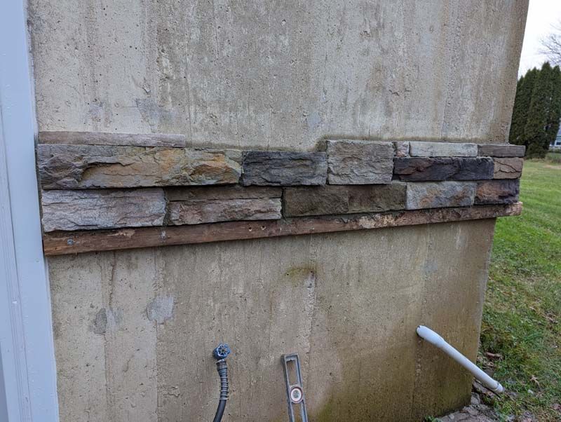 Stone veneer on a concrete wall, with pipes and grass in the background.