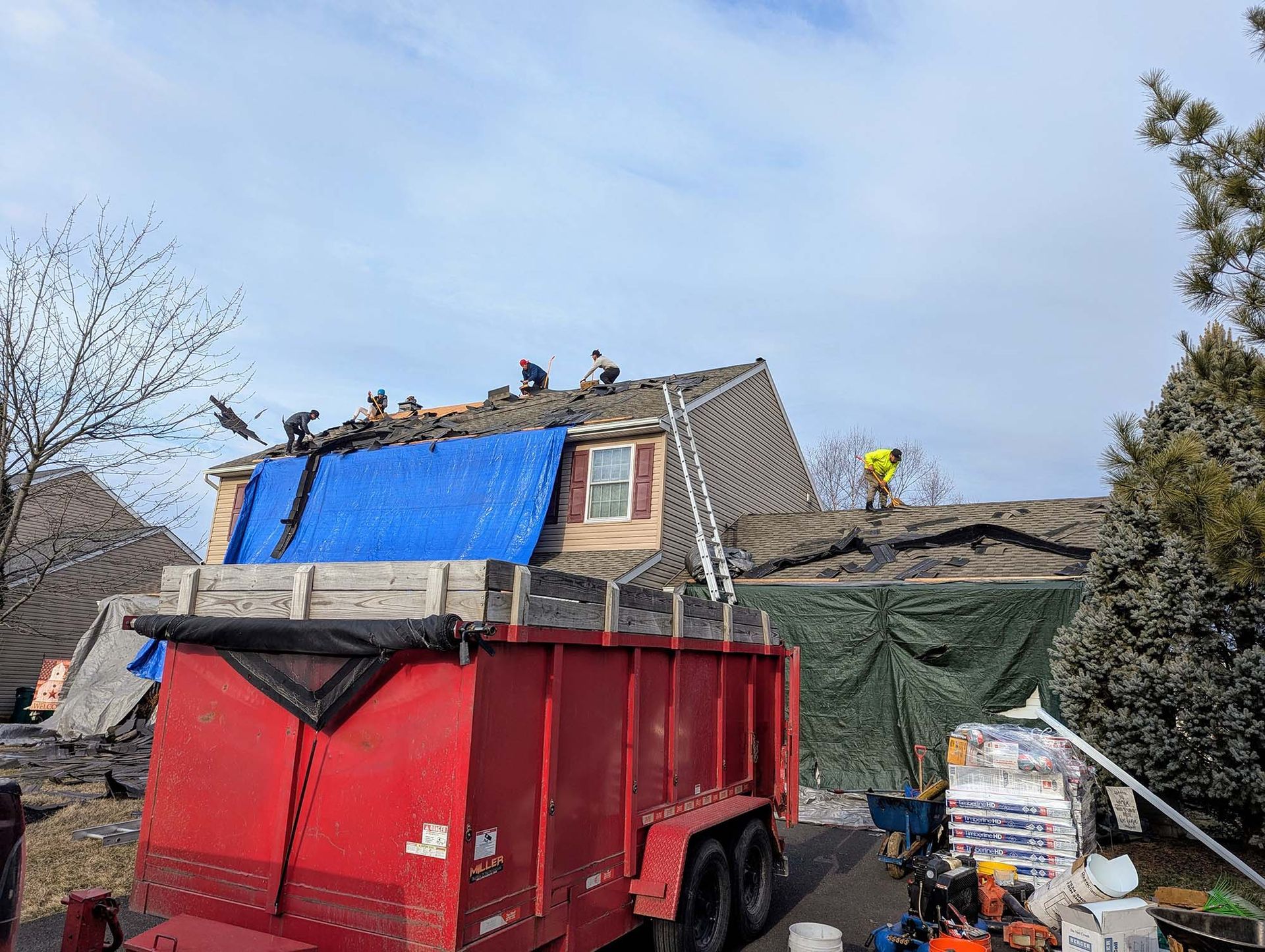 Roofers working on a house with a blue tarp, red dumpster, and green tarp. Sunny, outdoor setting.