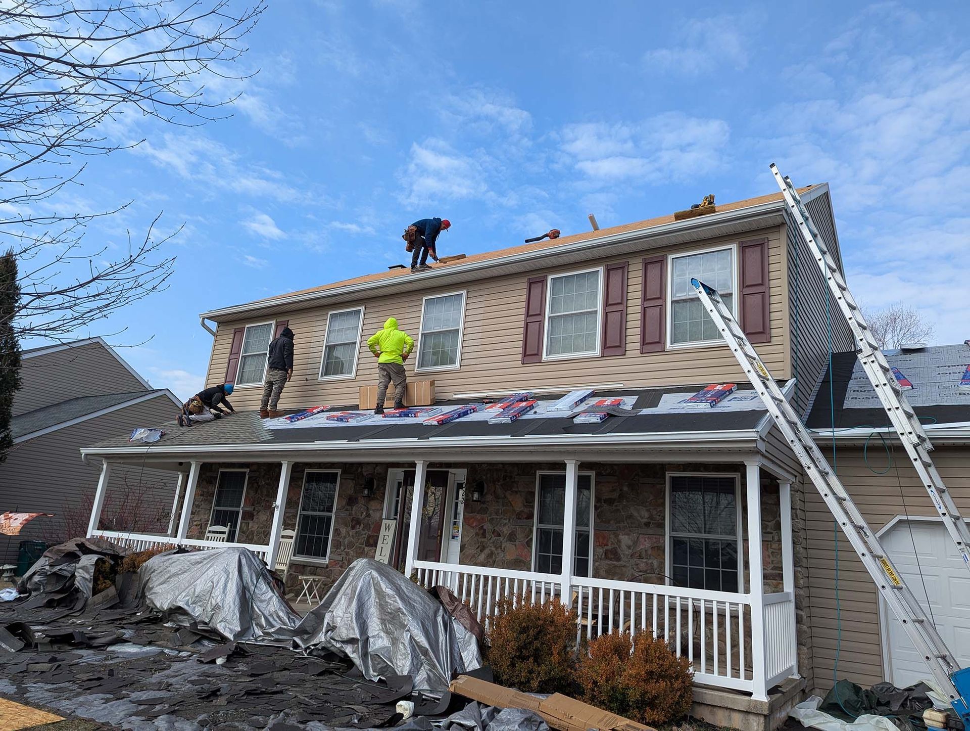 Roofers working on a two-story house with brown siding, ladders, and a blue sky.