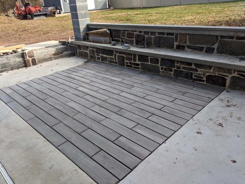Concrete patio with gray plank pavers. Stone wall with a concrete bench in the background.
