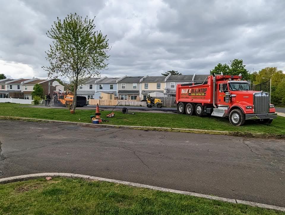Red dump truck parked near a road with asphalt paving in progress; cloudy sky and suburban homes in the background.